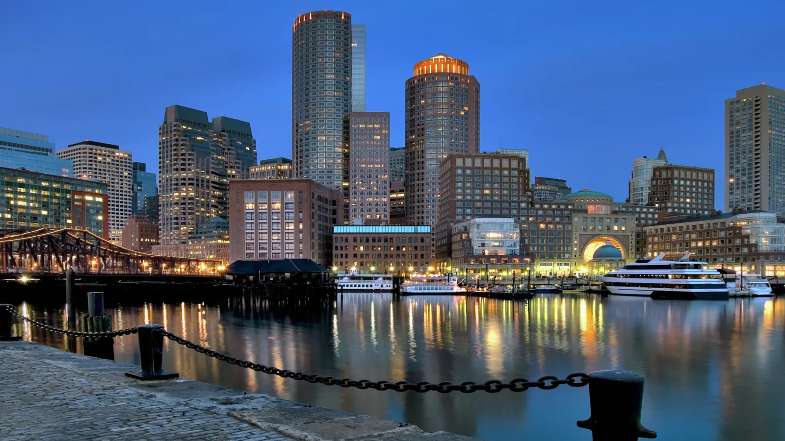A cityscape of tall buildings along a waterfront with boats docked and reflections in the water.