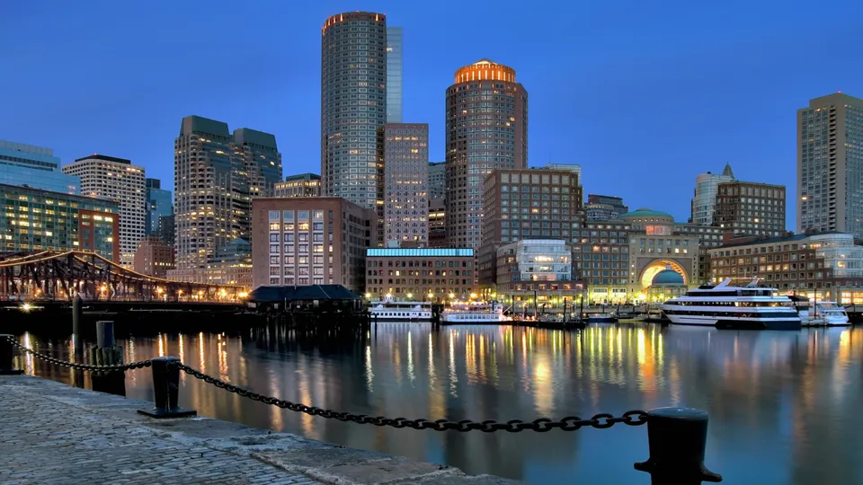 A cityscape of tall buildings along a waterfront with boats docked and reflections in the water.