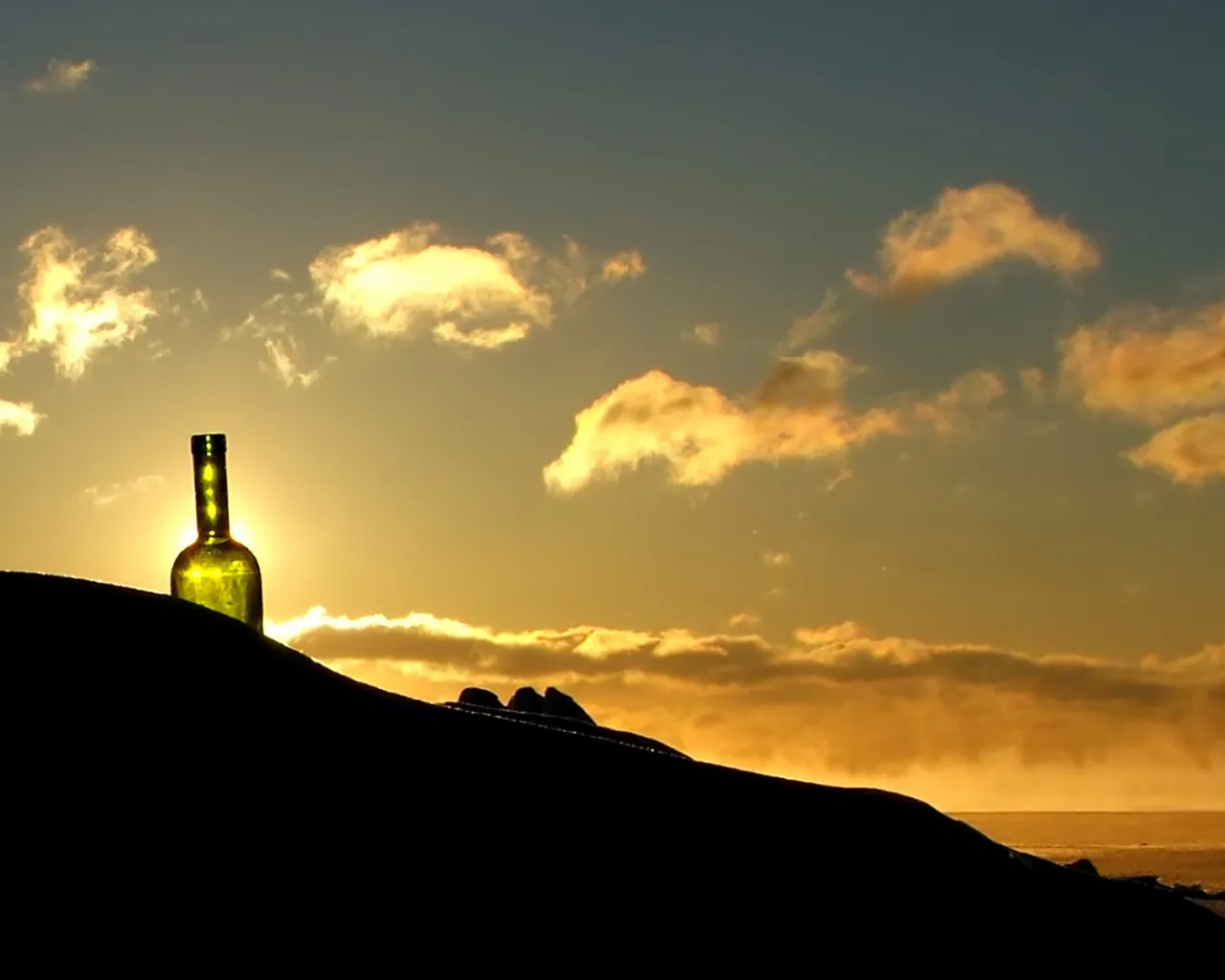 A green glass bottle is silhouetted against a golden sunset sky with scattered clouds.