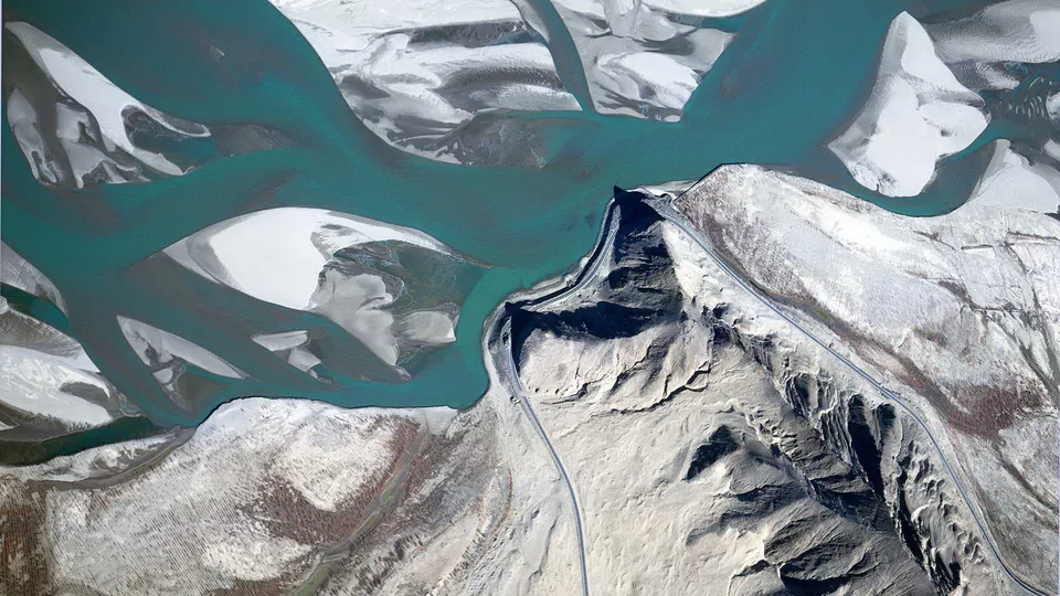 An aerial view shows a turquoise braided river flowing through a barren, mountainous landscape with patches of snow.