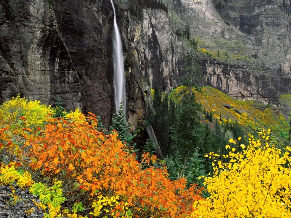 A tall waterfall cascades down a rocky cliff face, surrounded by vibrant autumn foliage.