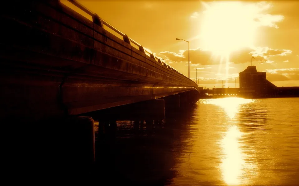 A long concrete bridge arches over a body of water, with the sun setting or rising and casting a golden glow.