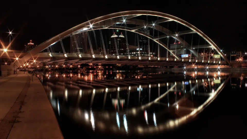 A modern suspension bridge illuminated at night, with its lights reflecting in the dark water below.