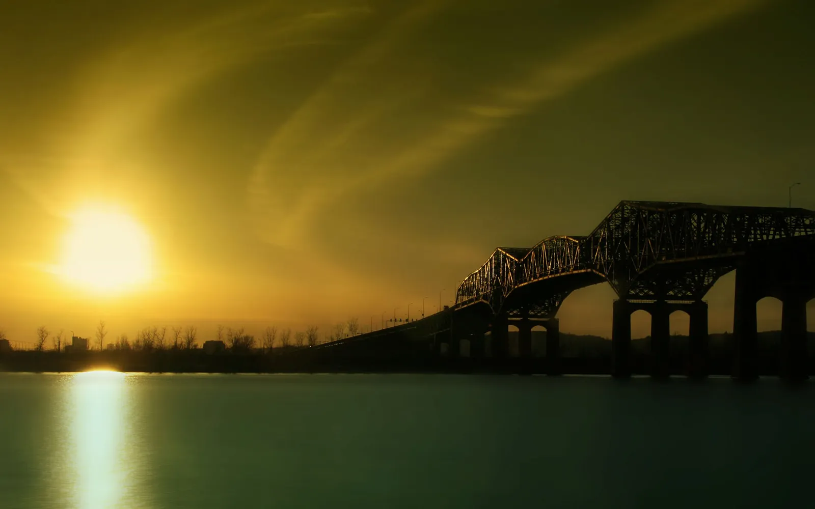 A large metal truss bridge spans a calm river during a golden sunset.