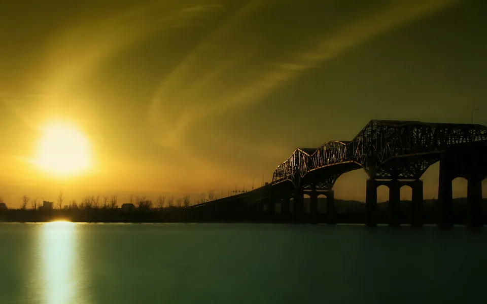 A large metal truss bridge spans a calm river during a golden sunset.