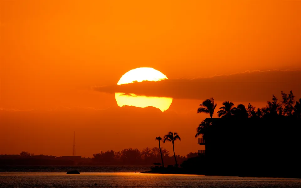A large, bright sun is partially obscured by a cloud during a vibrant orange sunset over the ocean.