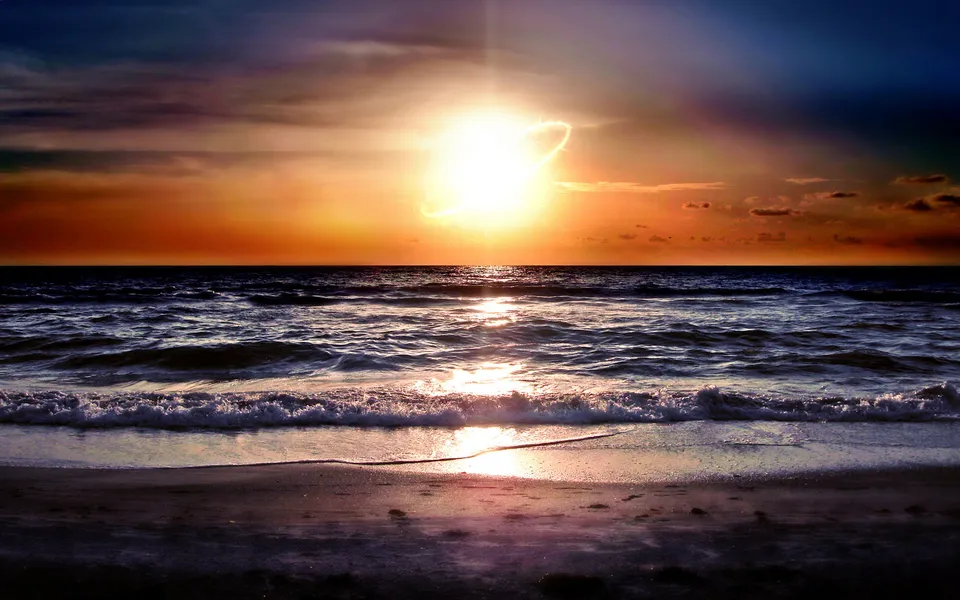 The sun sets over the ocean, casting bright light on the waves and beach.