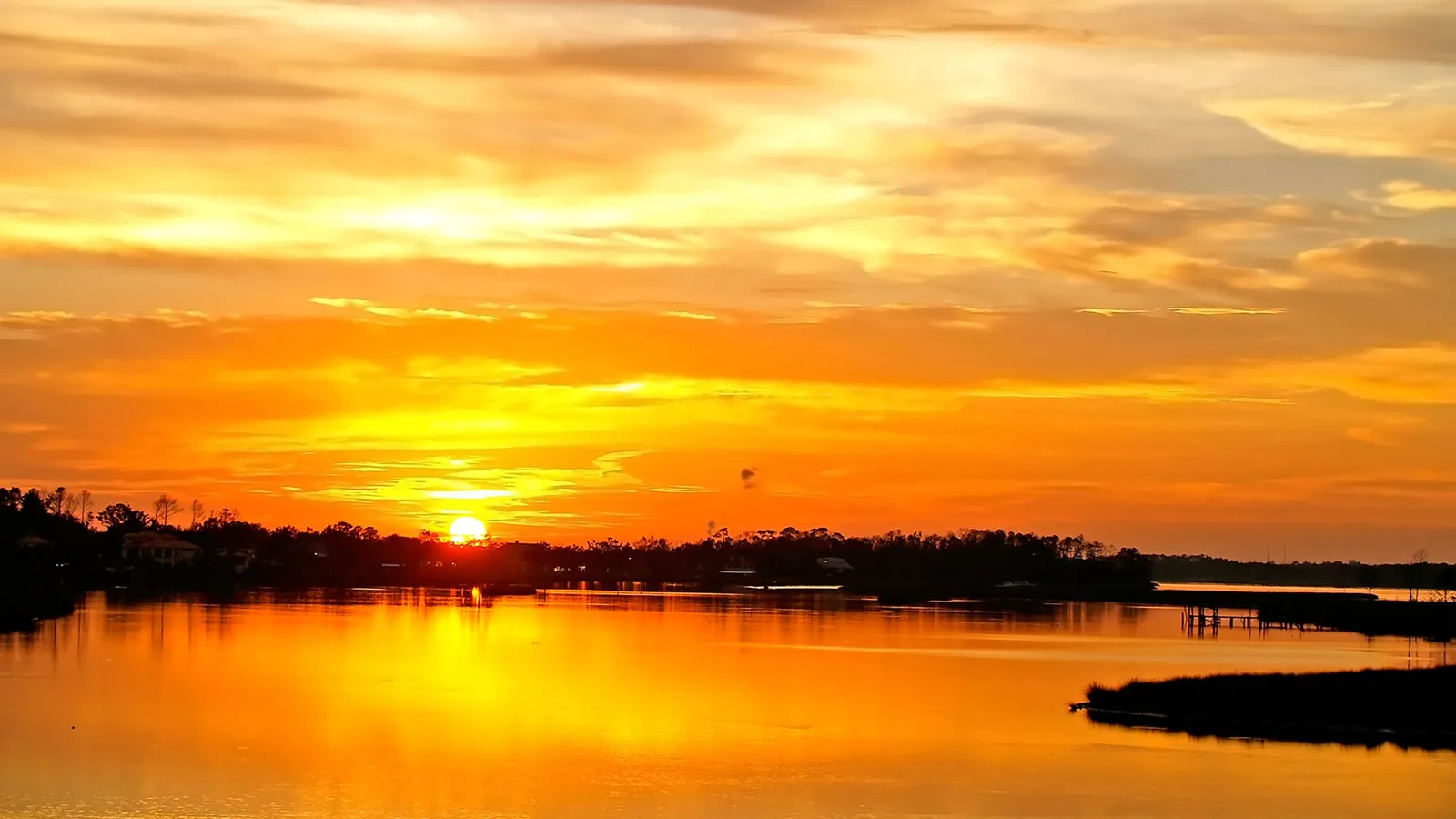 A brilliant orange sunset sky is reflected on the calm water of a coastal scene.