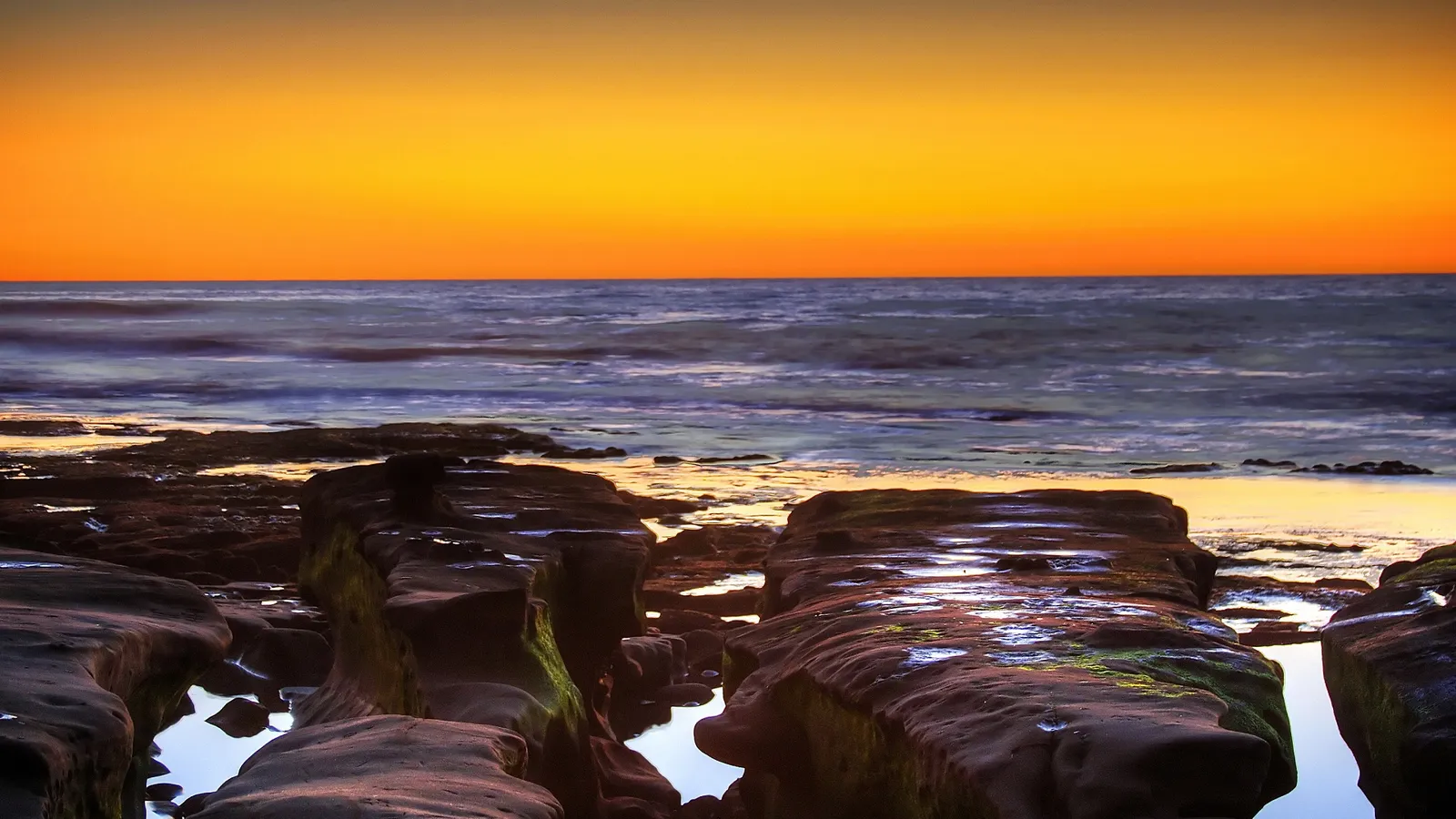 A vibrant orange sunset over a rocky coastline with tide pools reflecting the sky.