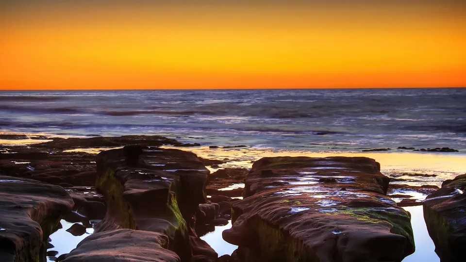 A vibrant orange sunset over a rocky coastline with tide pools reflecting the sky.