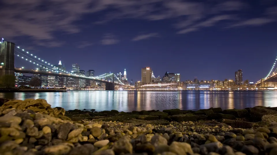 The Brooklyn Bridge and Manhattan skyline are illuminated at night, with reflections in the water.