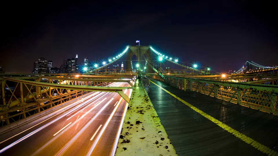 The Brooklyn Bridge at night, with car light trails on the roadway and the New York City skyline in the background.