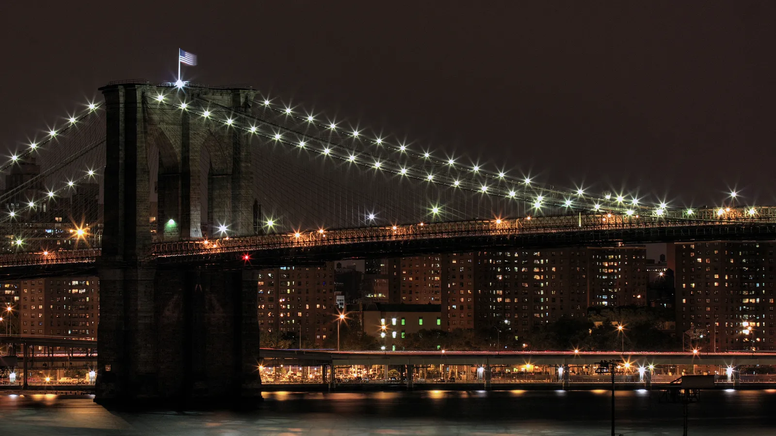 The Brooklyn Bridge is lit up at night with many lights, with city buildings in the background.