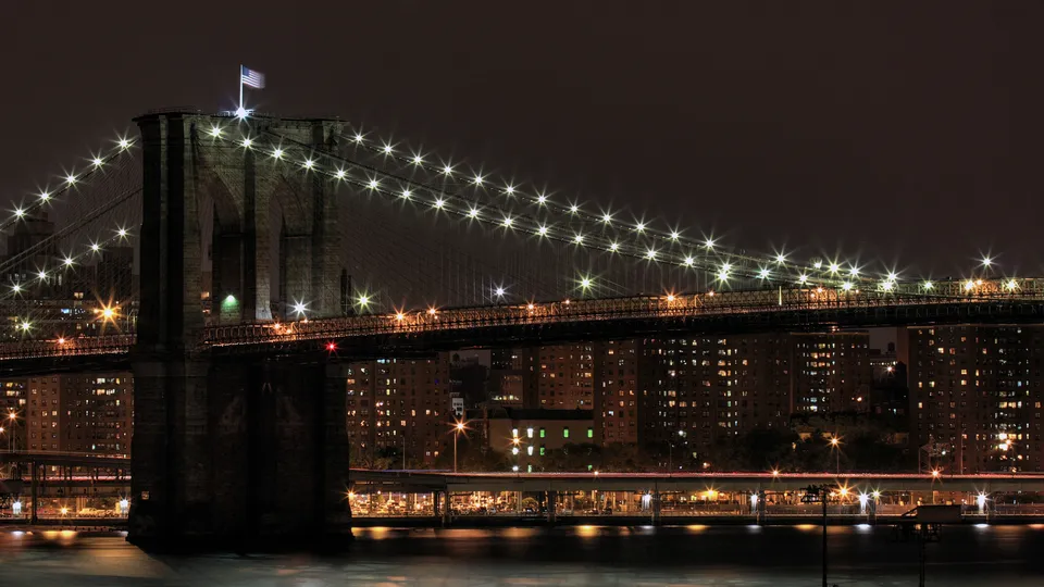 The Brooklyn Bridge is lit up at night with many lights, with city buildings in the background.