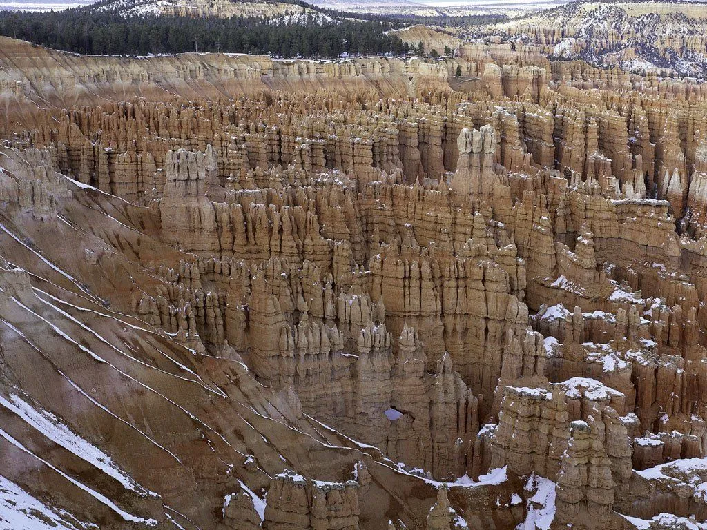 A vast landscape of tall, narrow rock formations called hoodoos, with patches of snow on the slopes and tops.