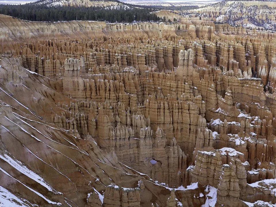 A vast landscape of tall, narrow rock formations called hoodoos, with patches of snow on the slopes and tops.
