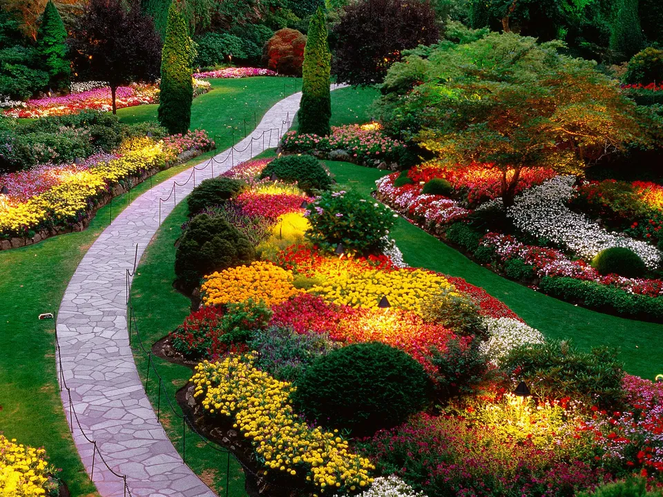 A stone pathway winds through a garden filled with colorful flowers and manicured bushes, illuminated by soft lighting.