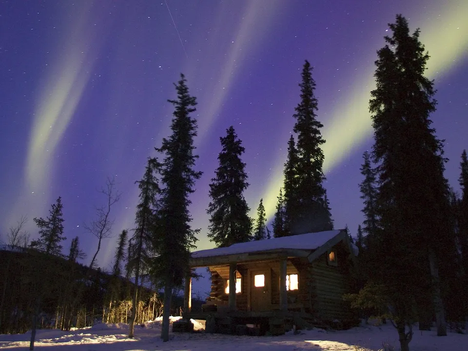 A small wooden cabin glows with warm light under a starry night sky with the aurora borealis dancing above.