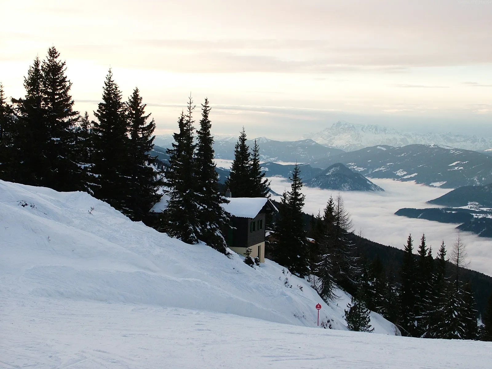 A small cabin sits on a snowy slope overlooking a valley filled with clouds.