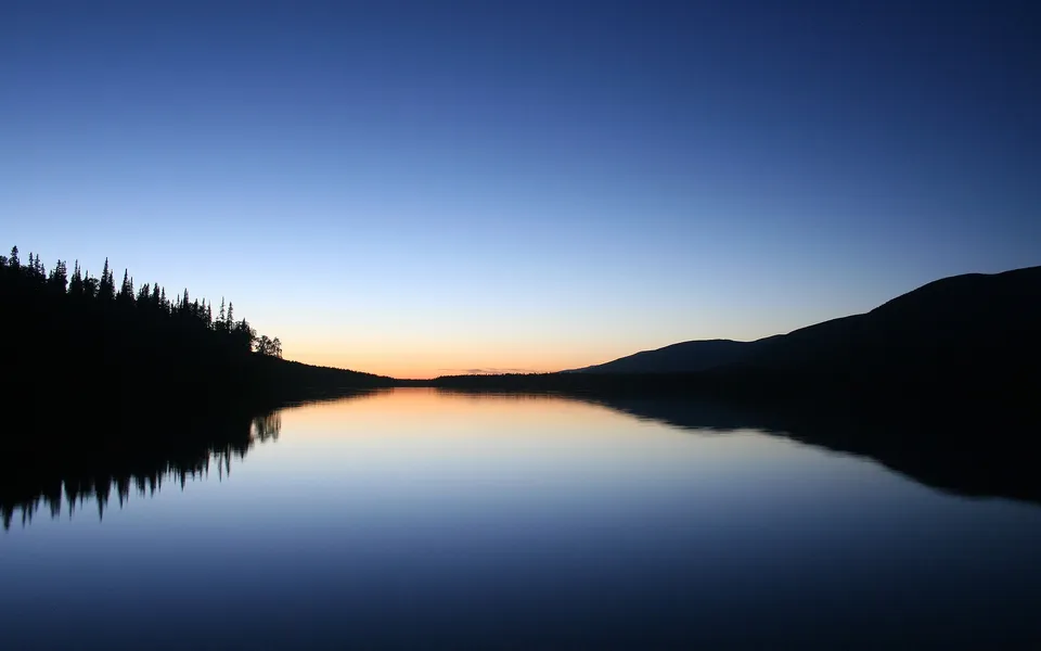 A calm lake reflects the deep blue and orange hues of the dusk sky, with silhouetted trees and hills on the horizon.