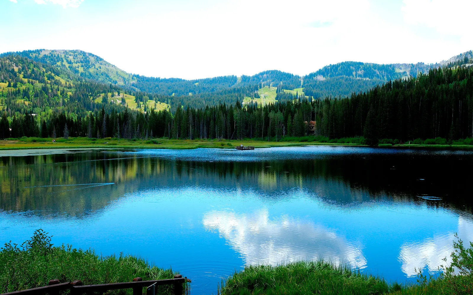 A calm lake perfectly mirrors the lush green mountains and a bright blue sky with white clouds.