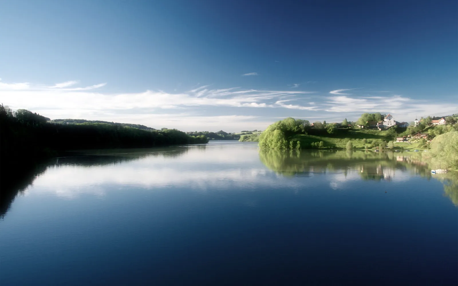 A wide, calm lake reflects the blue sky and surrounding green hills with a small village on the right.