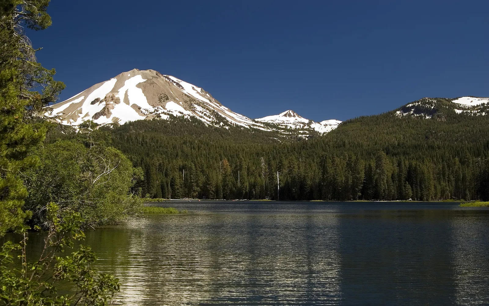 A tranquil lake with gentle ripples is in the foreground, with snow-dusted mountains and a dense evergreen forest behind it.