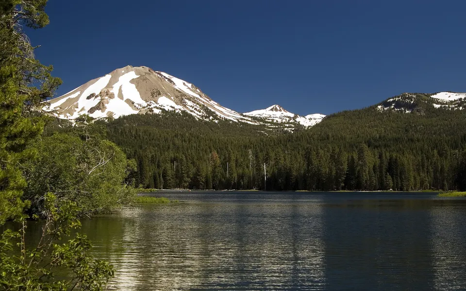 A tranquil lake with gentle ripples is in the foreground, with snow-dusted mountains and a dense evergreen forest behind it.