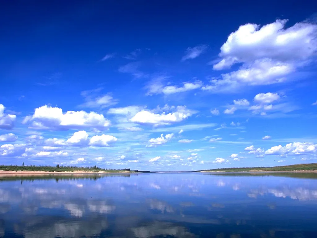 A wide, calm river perfectly reflects a bright blue sky filled with fluffy white clouds.