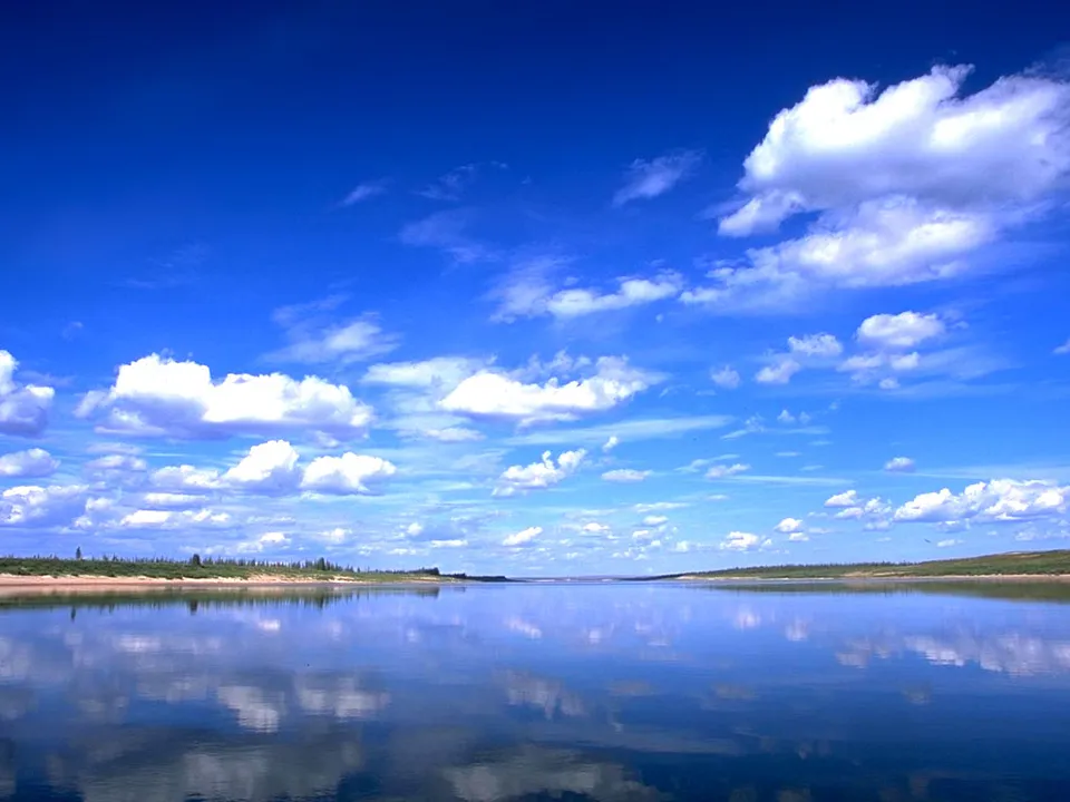 A wide, calm river perfectly reflects a bright blue sky filled with fluffy white clouds.