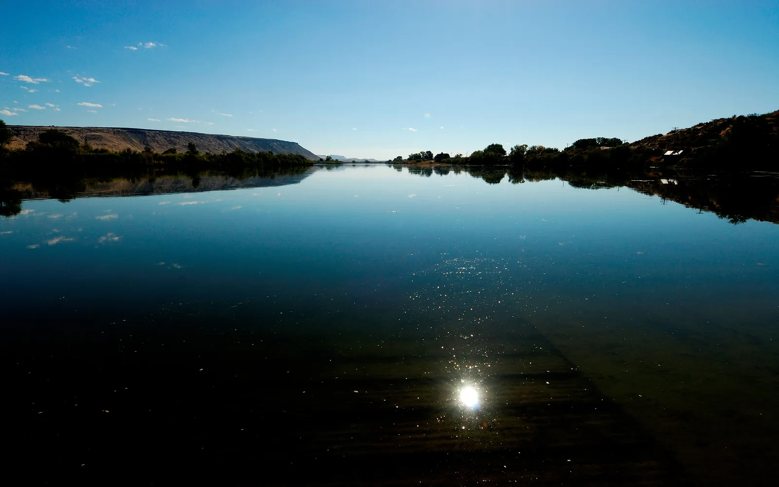 A wide, calm river reflects the clear blue sky and distant hills with trees.