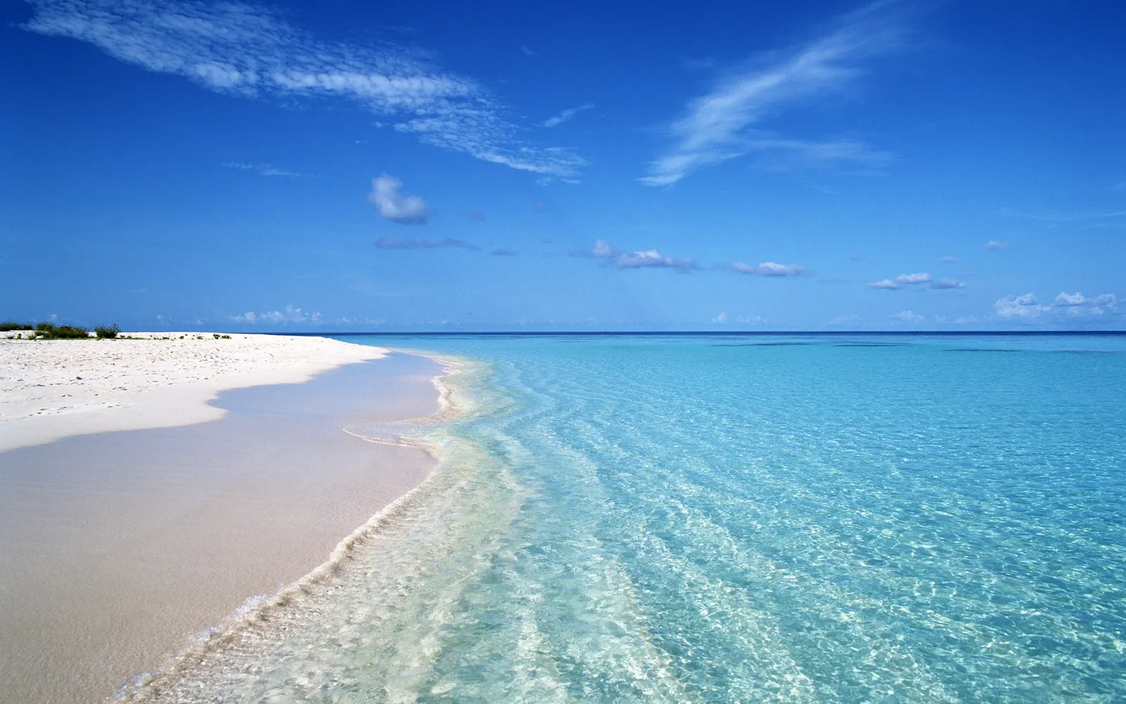 A serene tropical beach with fine white sand meeting calm, shallow turquoise waters under a bright blue sky.