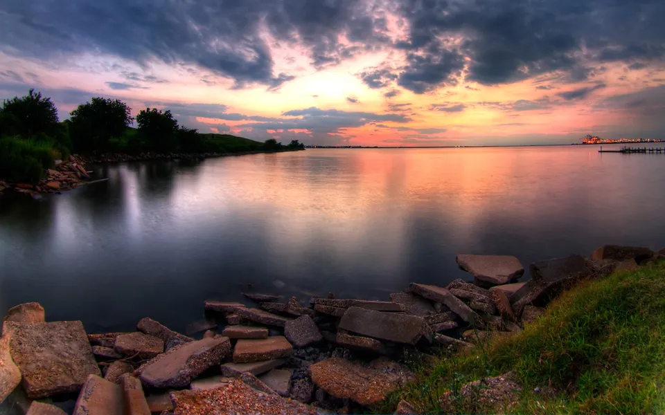 A dramatic sunset with colorful clouds over a calm body of water, with a rocky shore in the foreground.