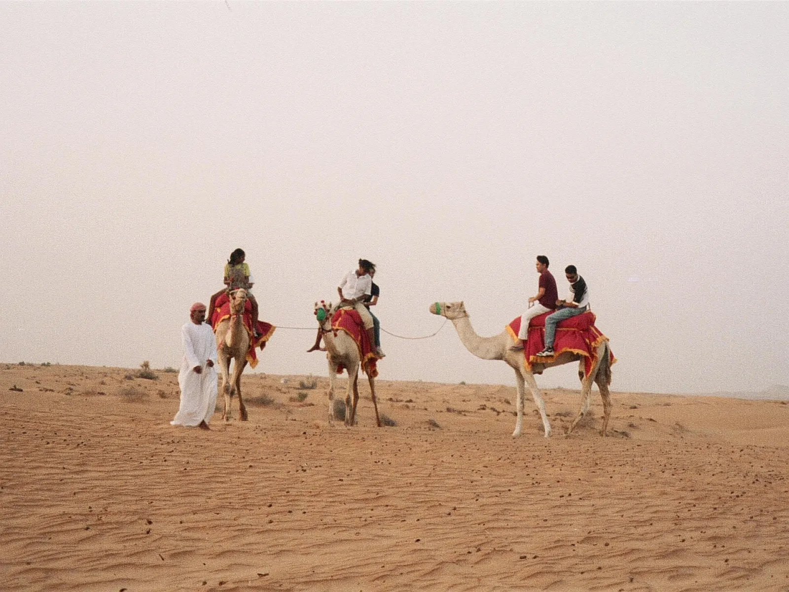 People riding camels across sand dunes under a hazy sky.