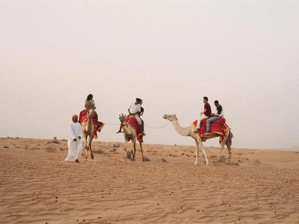 People riding camels across sand dunes under a hazy sky.