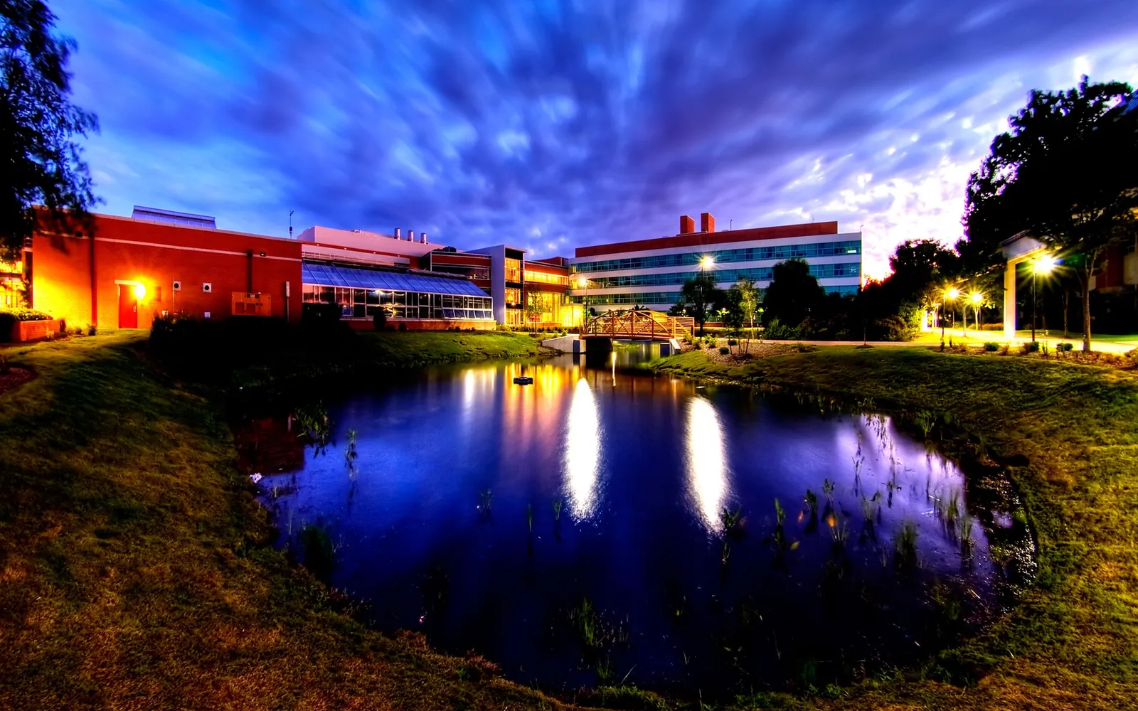 A modern building complex with lights on, reflected in a dark pond under a dramatic blue sky.