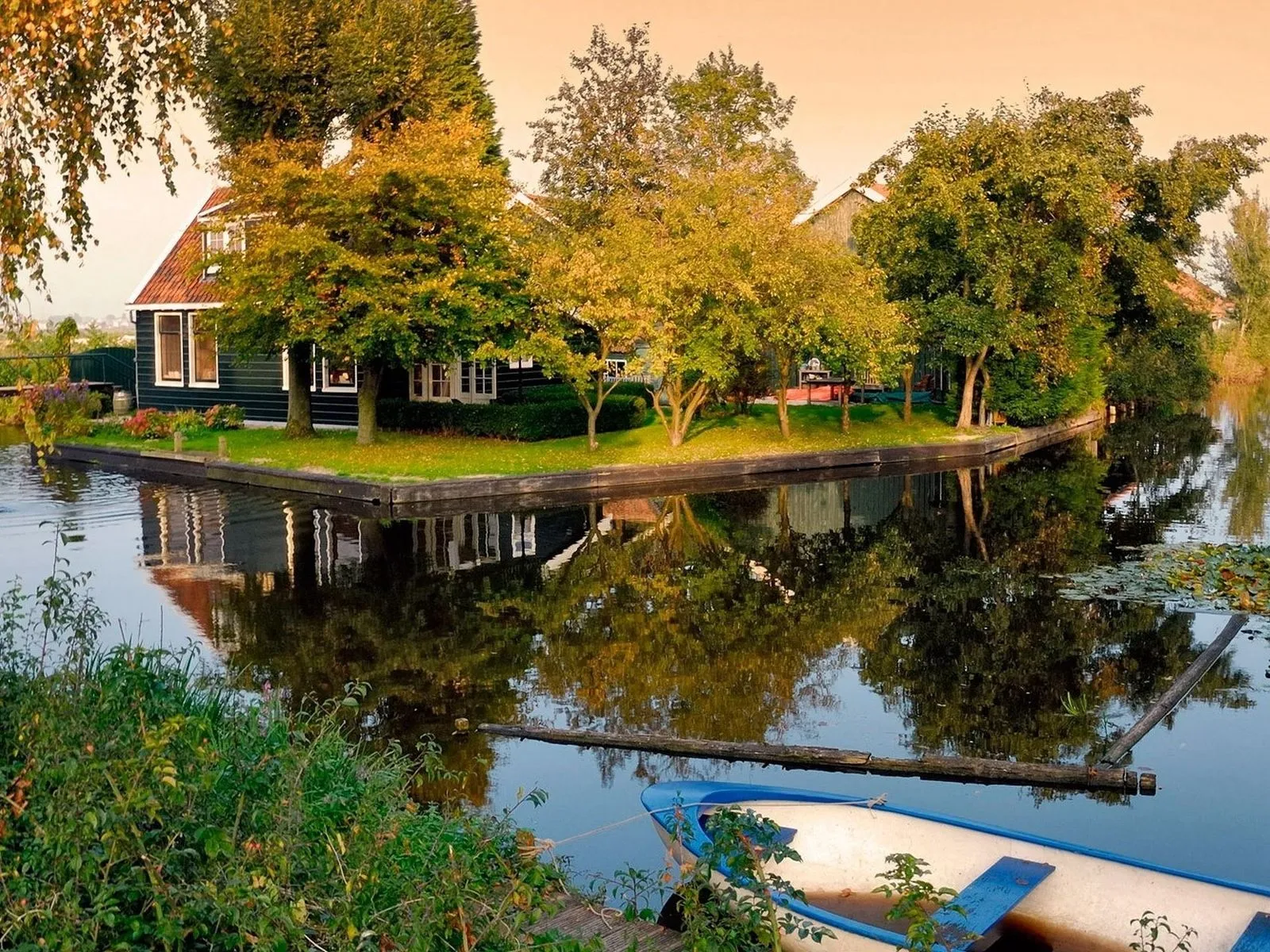 A dark green house with white trim sits on a narrow island surrounded by water, with trees and reflections.