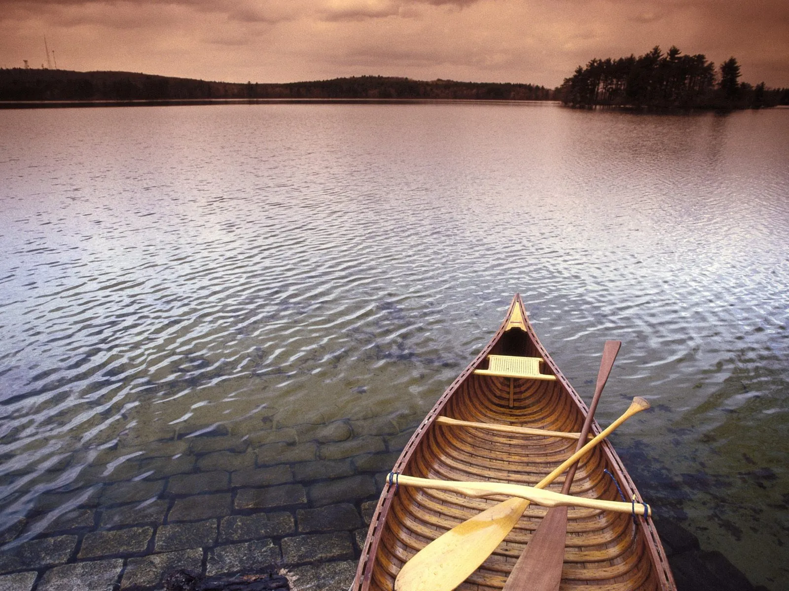 A wooden canoe with paddles rests on the edge of a calm lake under a cloudy sky.