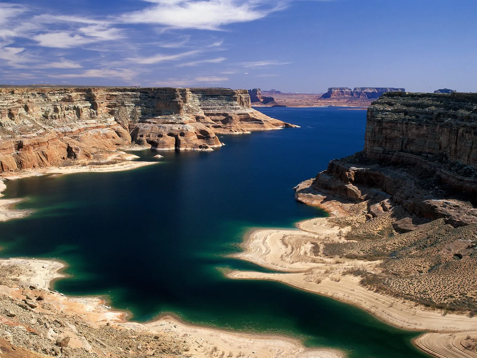 A wide, deep blue lake winds through towering sandstone canyons under a bright blue sky with scattered clouds.