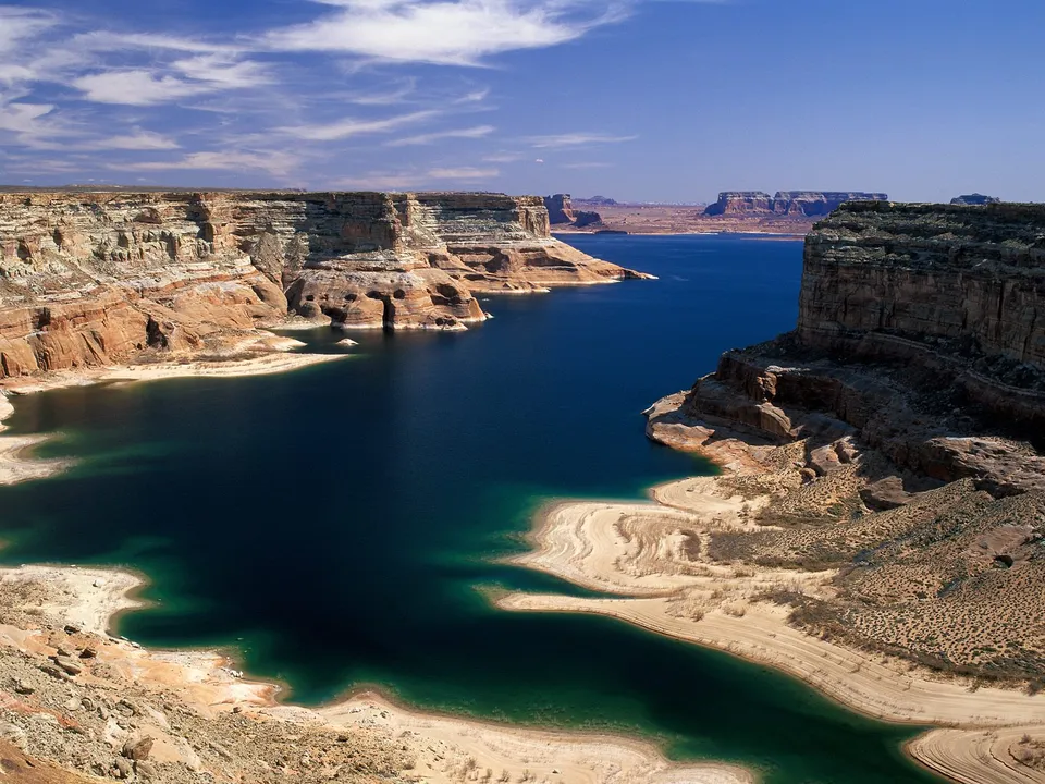 A wide, deep blue lake winds through towering sandstone canyons under a bright blue sky with scattered clouds.