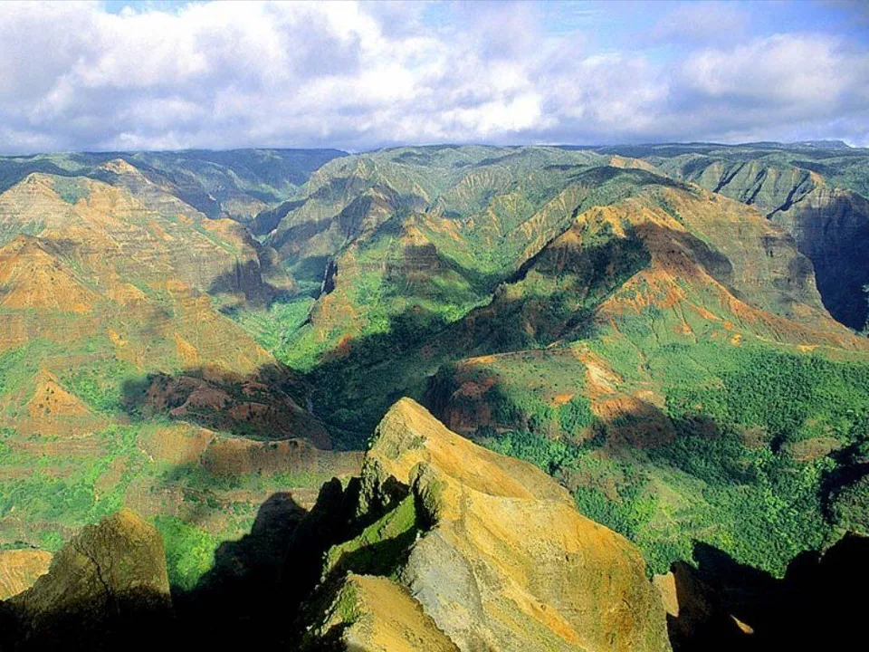 An aerial view of a vast, deep canyon with green vegetation and rocky slopes under a partly cloudy sky.