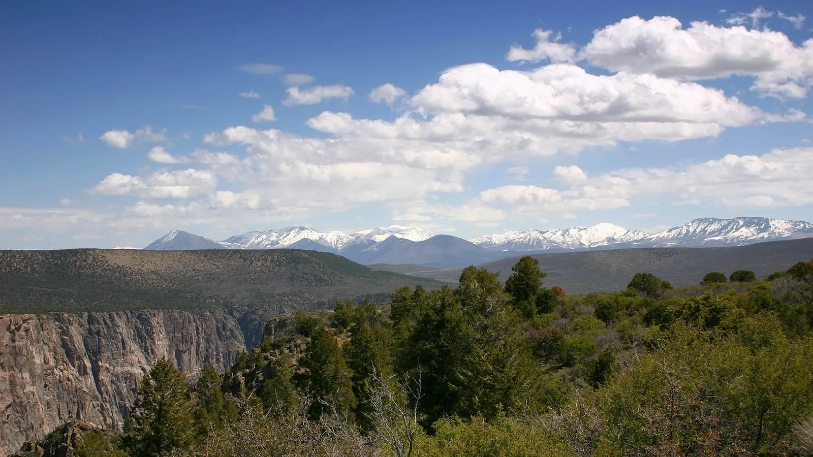 A wide view of a deep canyon with steep rocky walls, leading to a distant mountain range capped with snow under a blue sky with white clouds.