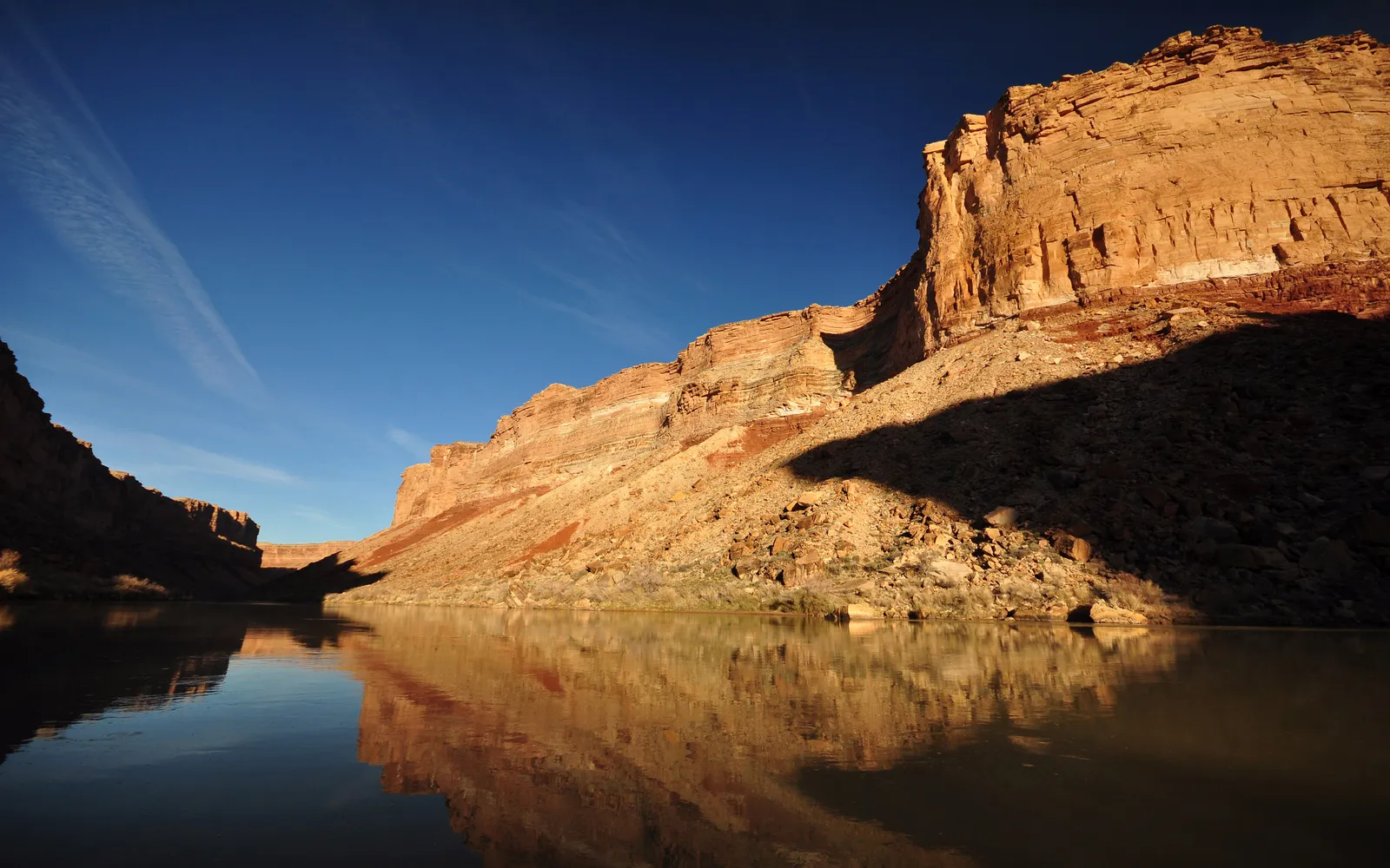 A wide, calm river flows through a canyon with steep, rocky, sunlit cliffs on the right and shadowed cliffs on the left.