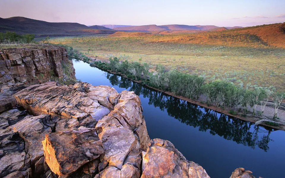 A wide river flows through a canyon with rocky cliffs in the foreground and rolling hills in the background at sunrise.