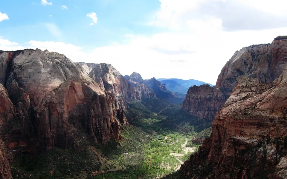 A wide view of a deep canyon with steep, rocky walls and a green valley floor below, under a partly cloudy sky.