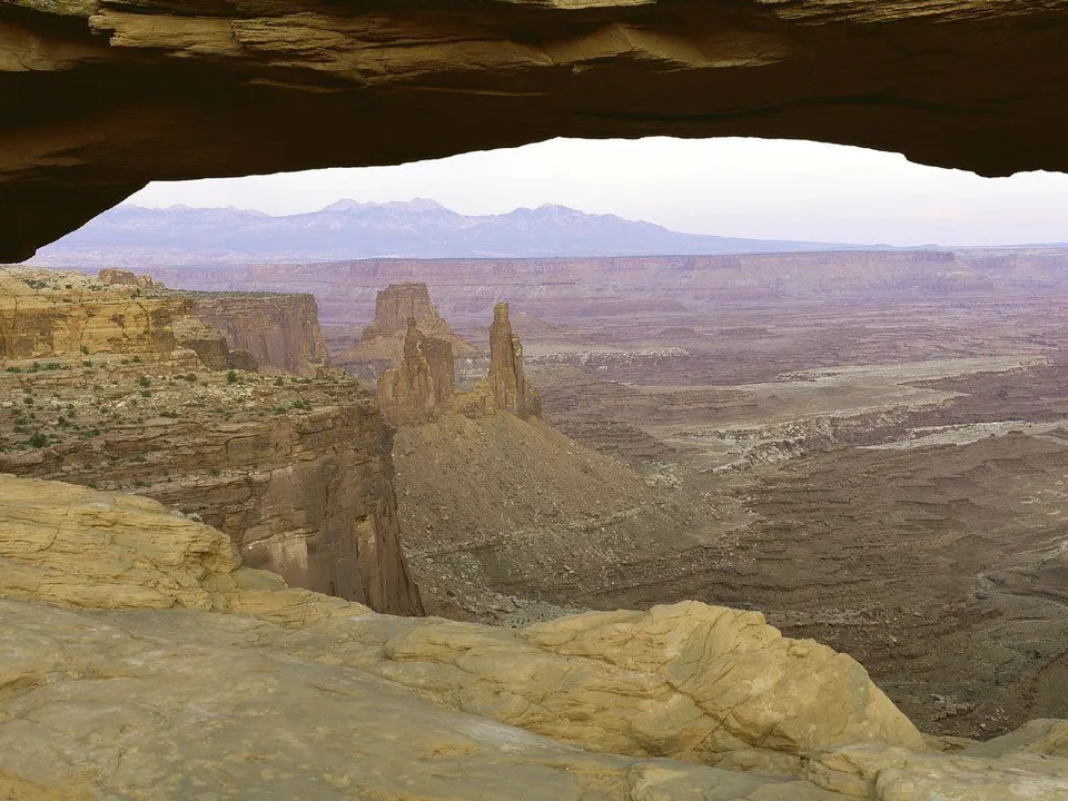 A wide desert canyon landscape viewed through a natural rock arch, with distant mountains on the horizon.