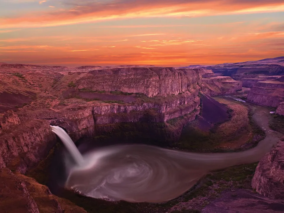 A wide waterfall cascades into a circular pool within a deep canyon at sunset.