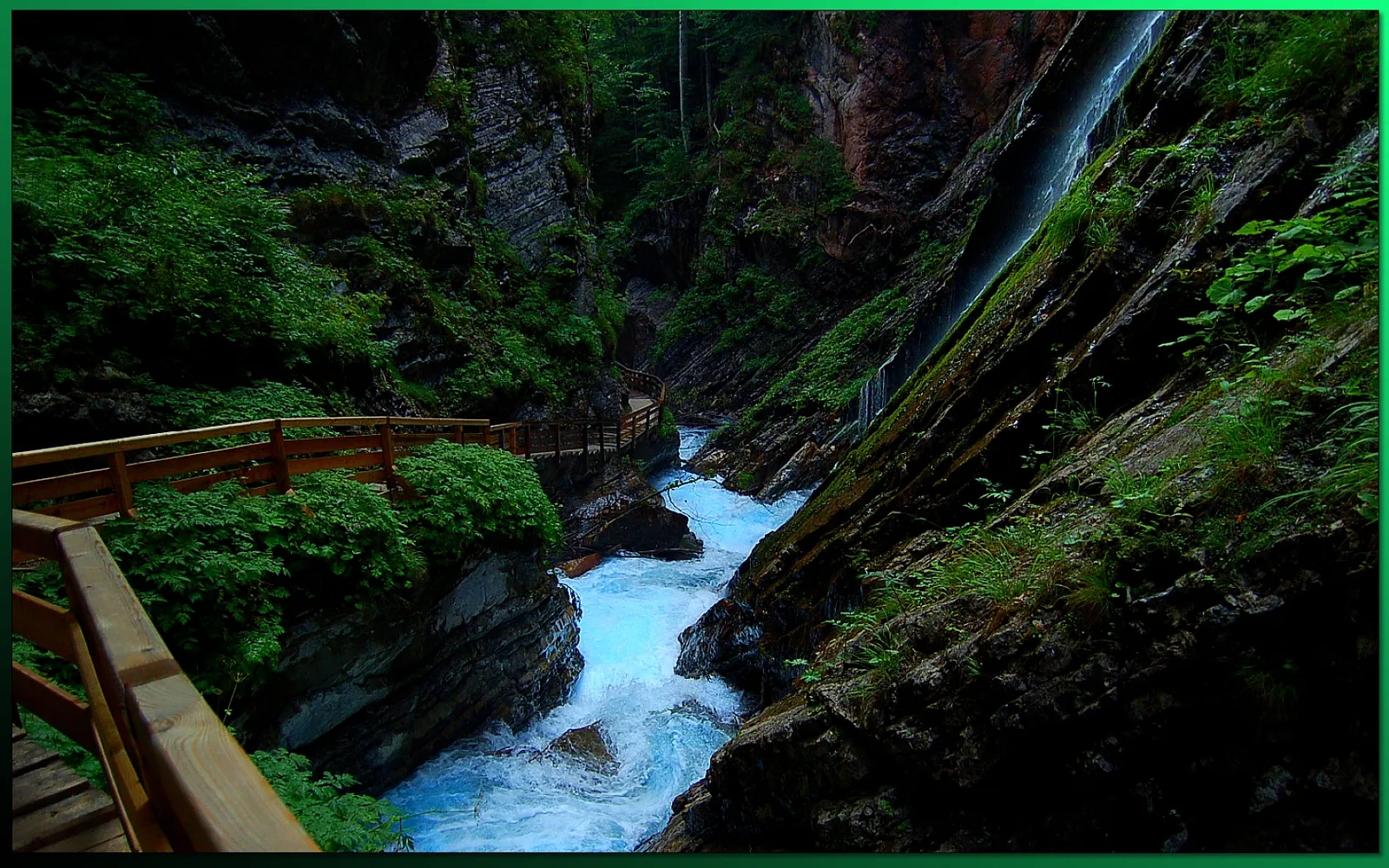 A wooden boardwalk follows a vibrant turquoise river through a deep, green canyon with a waterfall cascading down the rocks.