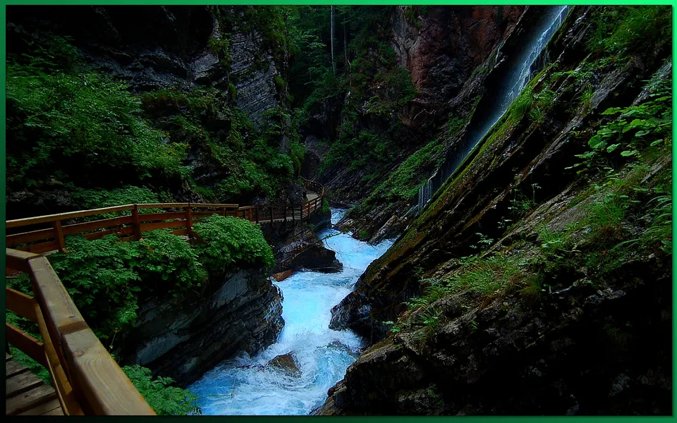 A wooden boardwalk follows a vibrant turquoise river through a deep, green canyon with a waterfall cascading down the rocks.