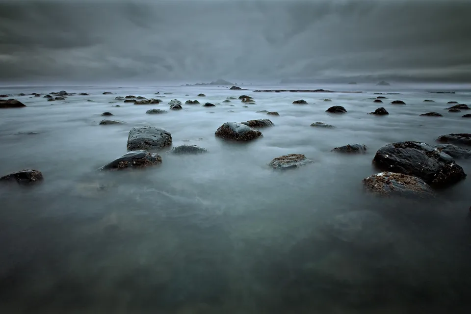 Smooth, misty water flows around dark, wet rocks on a cloudy day at the coast.